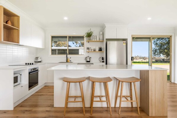 A modern kitchen with a central island, wooden stools, a built-in oven, and a view through large windows.