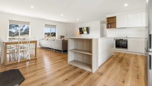 Una cocina moderna con armarios blancos, horno empotrado y comedor con mesa y sillas de madera.