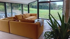A modern living room with a large sofa, a glass coffee table, and a view of a grassy outdoor area through large windows.
