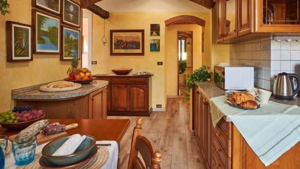 Une cuisine avec des meubles de rangement en bois, une table à manger sur laquelle se trouve un saladier rempli de fruits, et une machine à café.