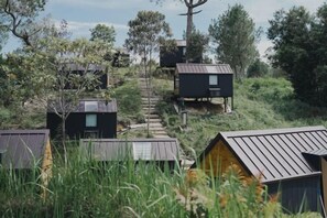A series of modern, dark-colored cabins with metal roofs, surrounded by lush greenery and trees. A wooden staircase leads up to the cabins.