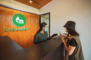 A reception desk with a wooden panel and a logo, a person standing behind it, and a counter. - Bobocabin Kaldera, Toba (Ajibata)