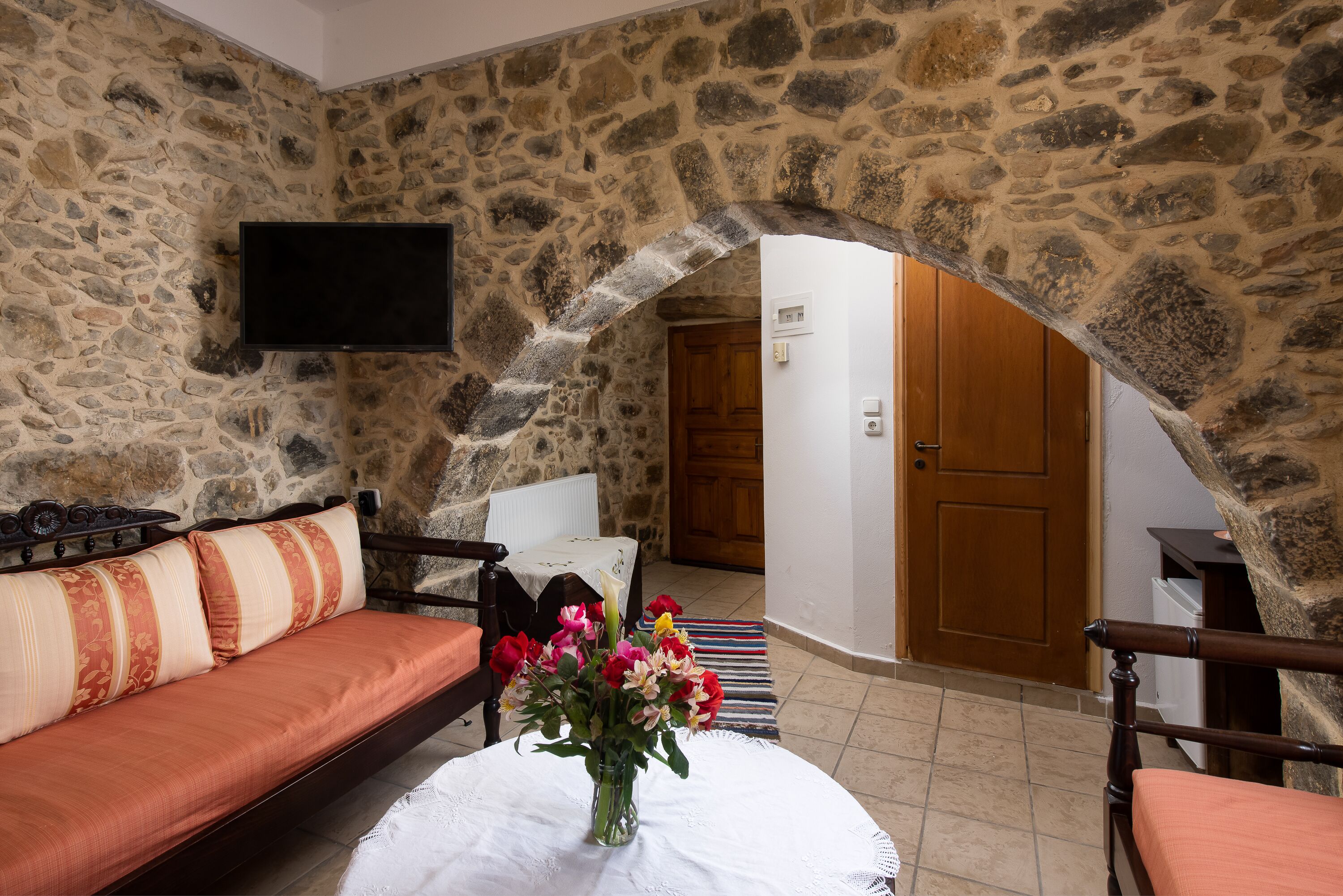 A stone-walled room with a sofa, a table with a vase of flowers, and a television mounted on the wall.