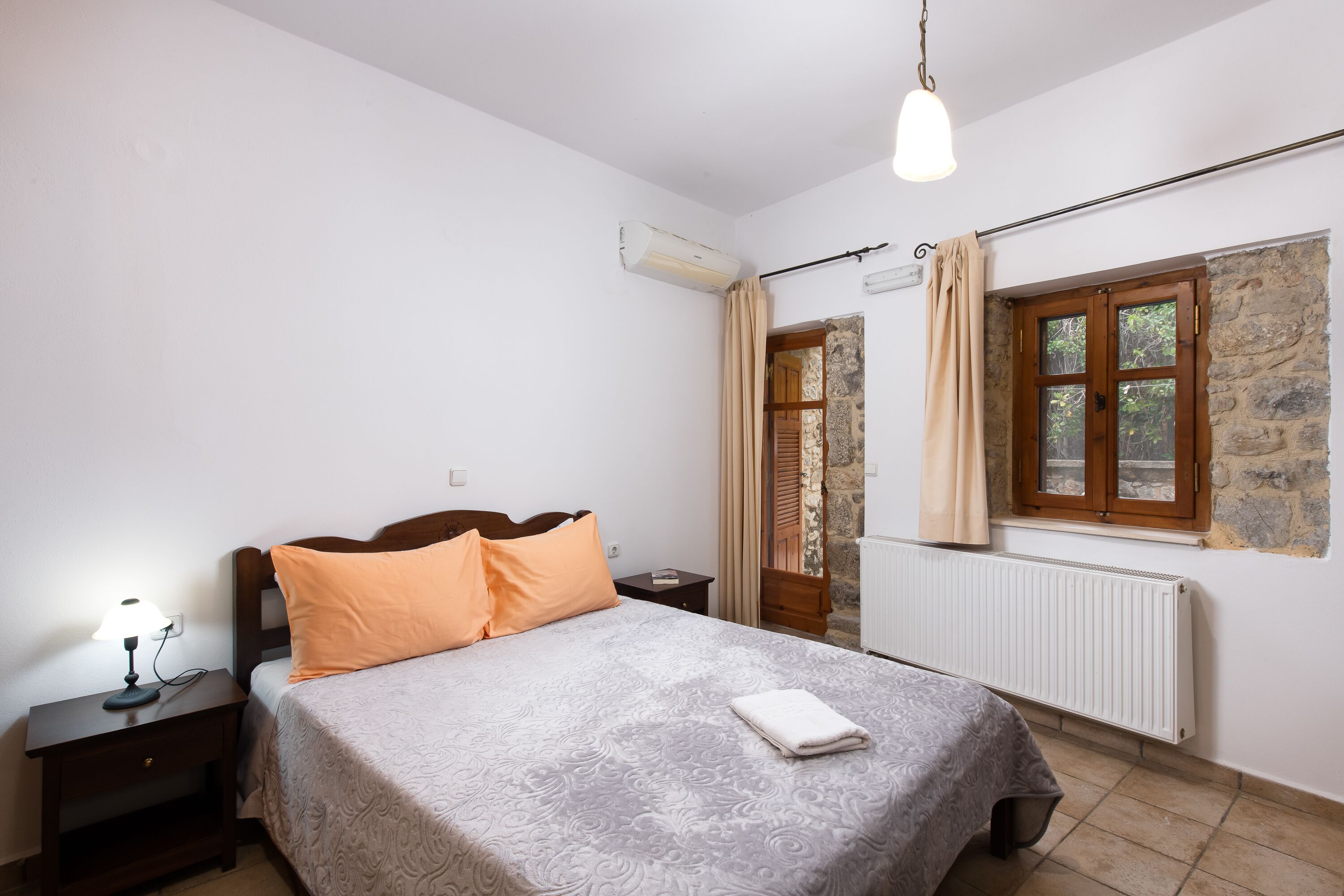 Quarto com cama, mesa de cabeceira, luminária, aparelho de ar condicionado, janela com cortinas e radiador.