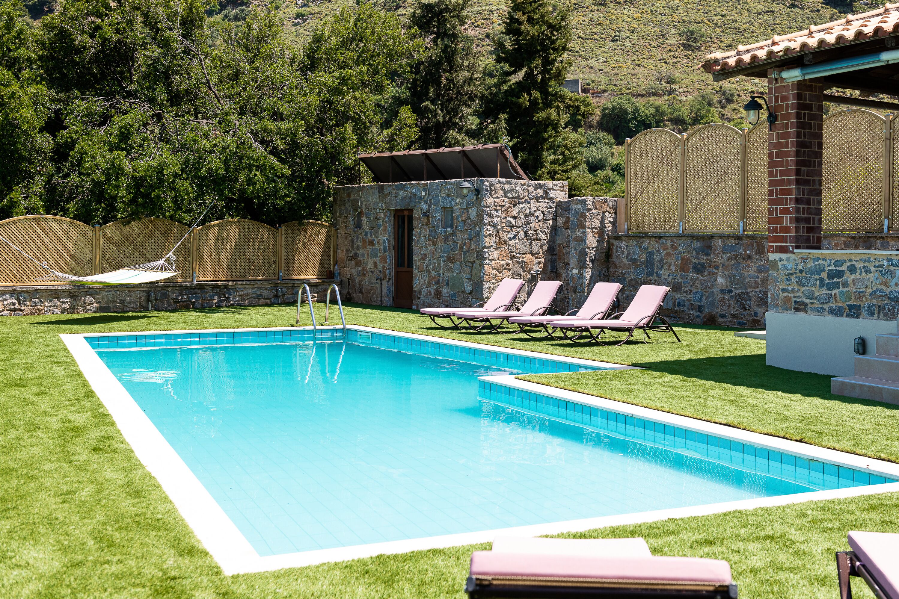 A rectangular swimming pool with a clear blue water, surrounded by a stone wall and a wooden fence. There are lounge chairs and a hammock near the pool.