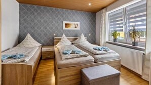 Ein Zimmer mit zwei Einzelbetten, einer Holzbalkendecke, einer grauen Wand mit Muster und einem Fenster mit Jalousien sowie einem Heizkörper.