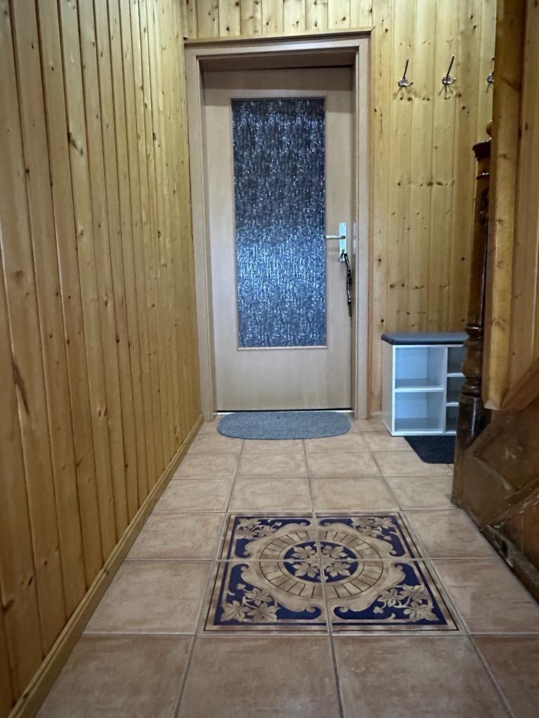 A hallway with wooden walls, a tiled floor with a decorative border, a door with a frosted glass panel, and a small white shelf unit.