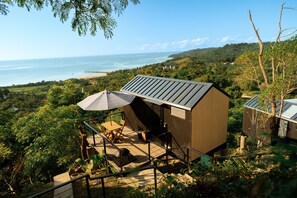A small wooden cabin with a sloped roof, a patio with a table and chairs, and a view of the ocean and hills. - Bobocabin Umarato Sumba (Wanokaka)
