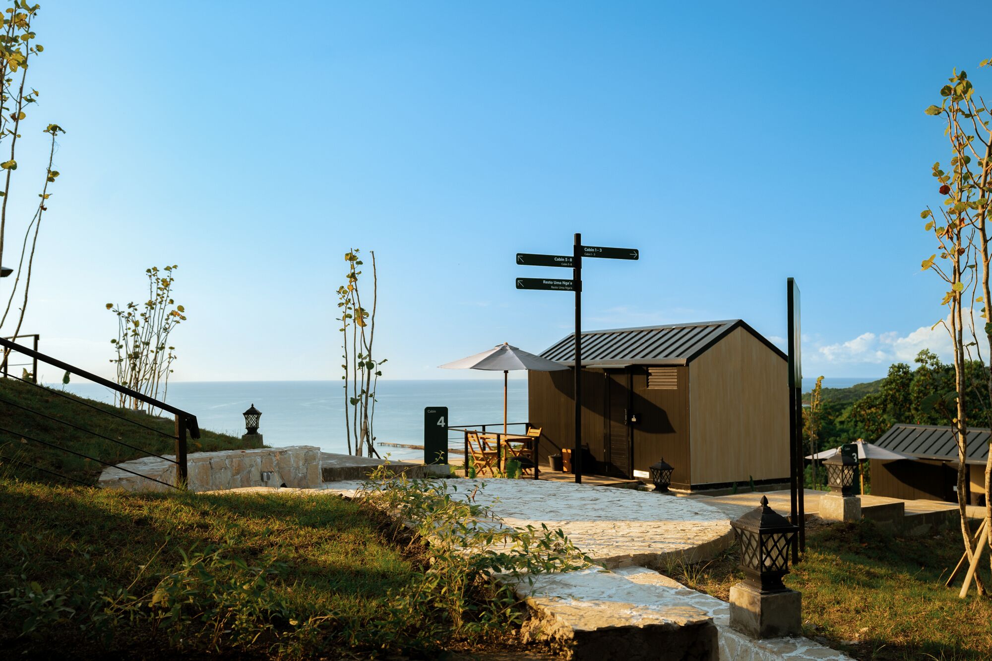 A coastal building with a stone pathway leading to a wooden structure, a signpost with directions, and a view of the sea.