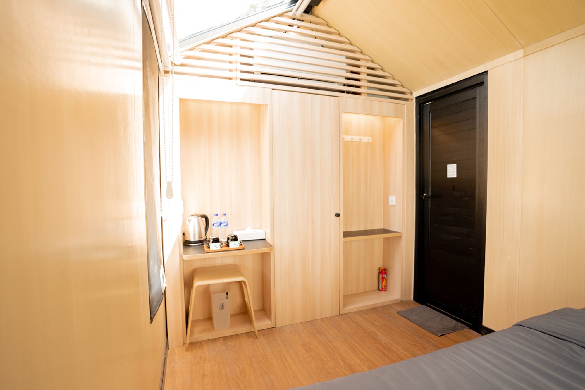 Una habitación pequeña y bien iluminada, con un escritorio de madera, una cama y una puerta negra.