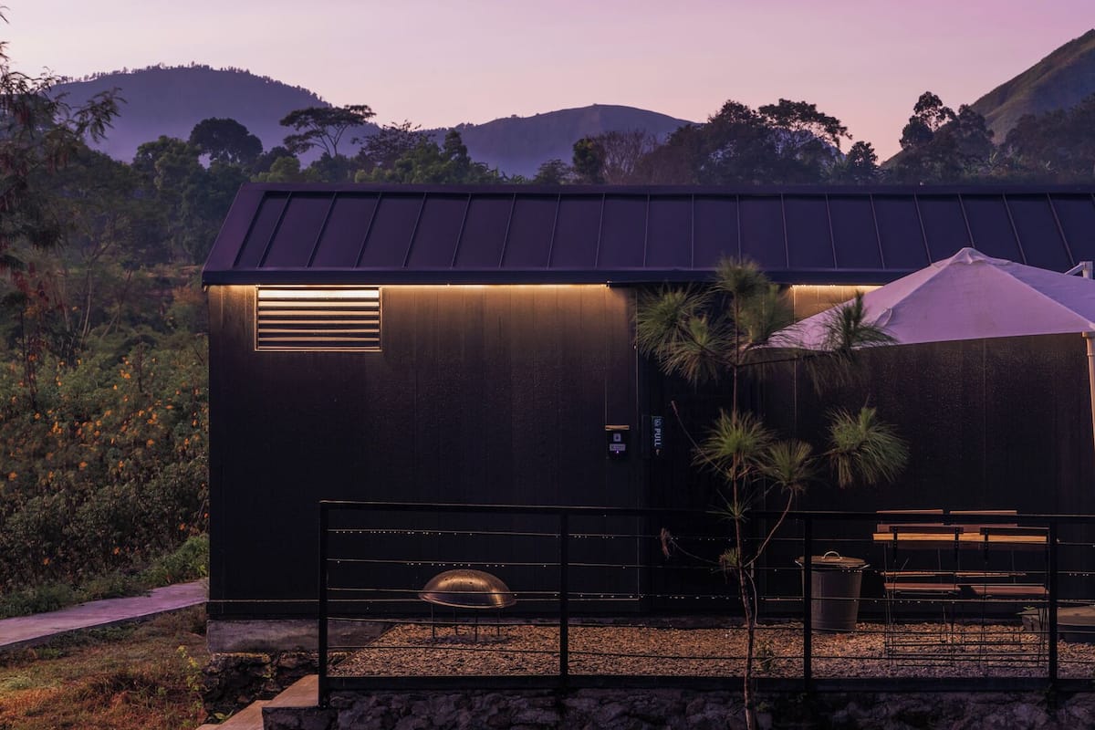 Un edificio moderno con techo metálico, un pequeño espacio al aire libre con mesas y sillas, y un camino de grava que lleva a la entrada.