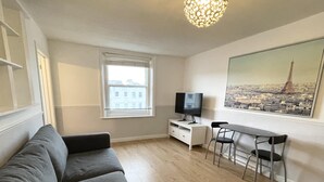 Un salon avec un canapé gris, un téléviseur posé sur un meuble blanc, une table à manger avec deux chaises, une fenêtre donnant sur des immeubles et un tableau représentant un paysage urbain.