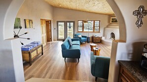 Un soggiorno spazioso con soffitto in legno a vista, un camino e vista sull'esterno attraverso ampie finestre.