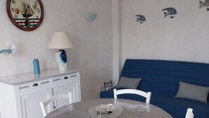 A dining area with a round table, white chairs, a blue sofa, a white cabinet, a decorative clock, and wall art. - Charming apartment with WIFI 5 minutes walk from the beach and town center (Saint-Palais-sur-Mer)