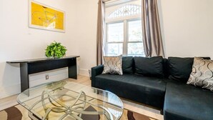 A modern living room with a glass coffee table, a black sofa with patterned cushions, a black console table with a plant, and a large window with curtains. - Beach House Oasis - 2BR Apartment with backyard patio in the Beaches Area (Toronto)