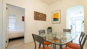 A dining area with a glass table and chairs, a bed in the adjacent room, and a framed picture on the wall. - Beach House Oasis - 2BR Apartment with backyard patio in the Beaches Area (Toronto)