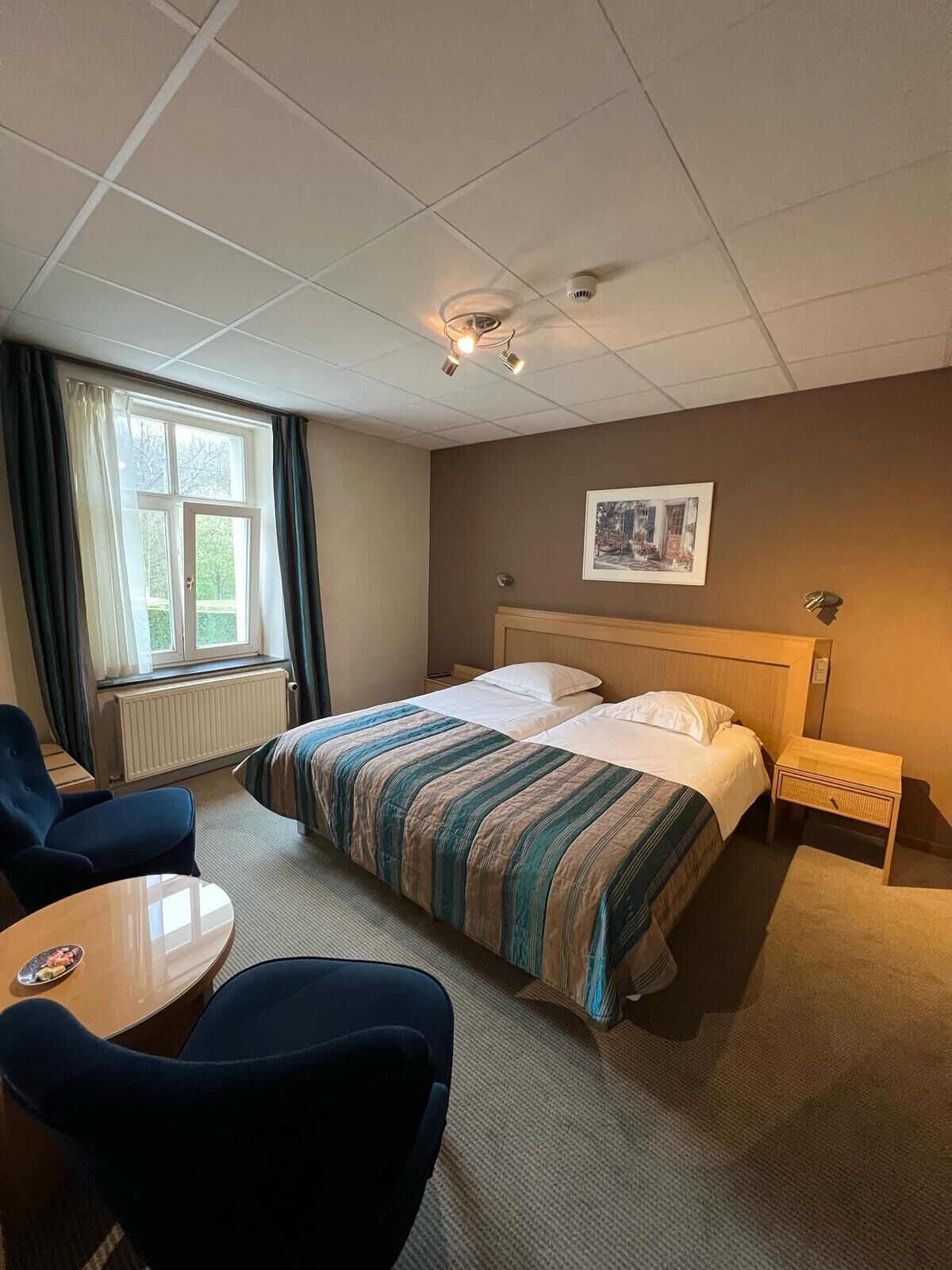 Une chambre d’hôtel avec un lit, un bureau, une chaise, une fenêtre avec des rideaux et un luminaire au plafond.
