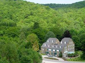 Un edificio grande de dos pisos con un techo simétrico y numerosas ventanas, rodeado por un bosque verde denso.
