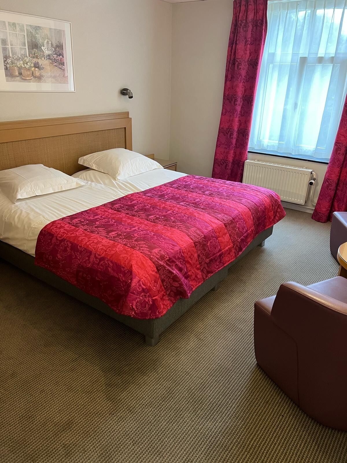 Une chambre d’hôtel avec un lit, une chaise, un radiateur et une fenêtre avec des rideaux.