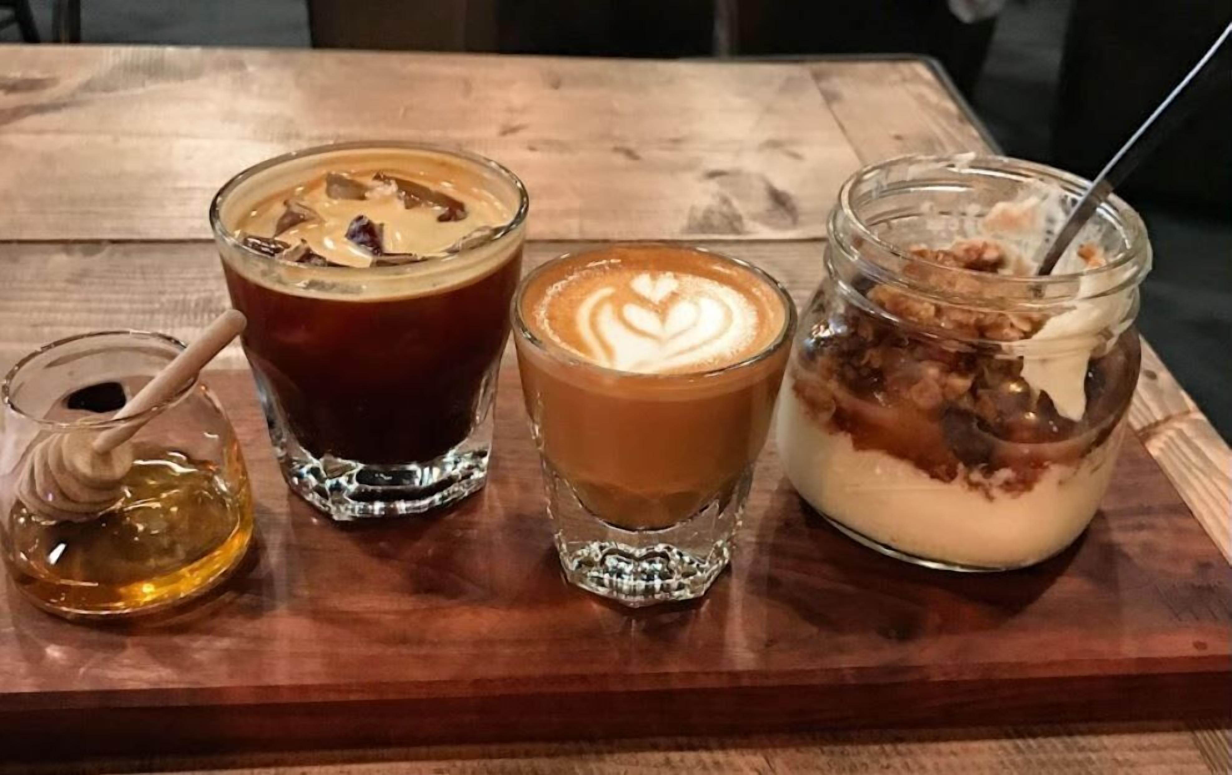 Three drinks on a wooden table: a dark beverage with ice cubes, a latte with latte art, and a jar with a spoon.