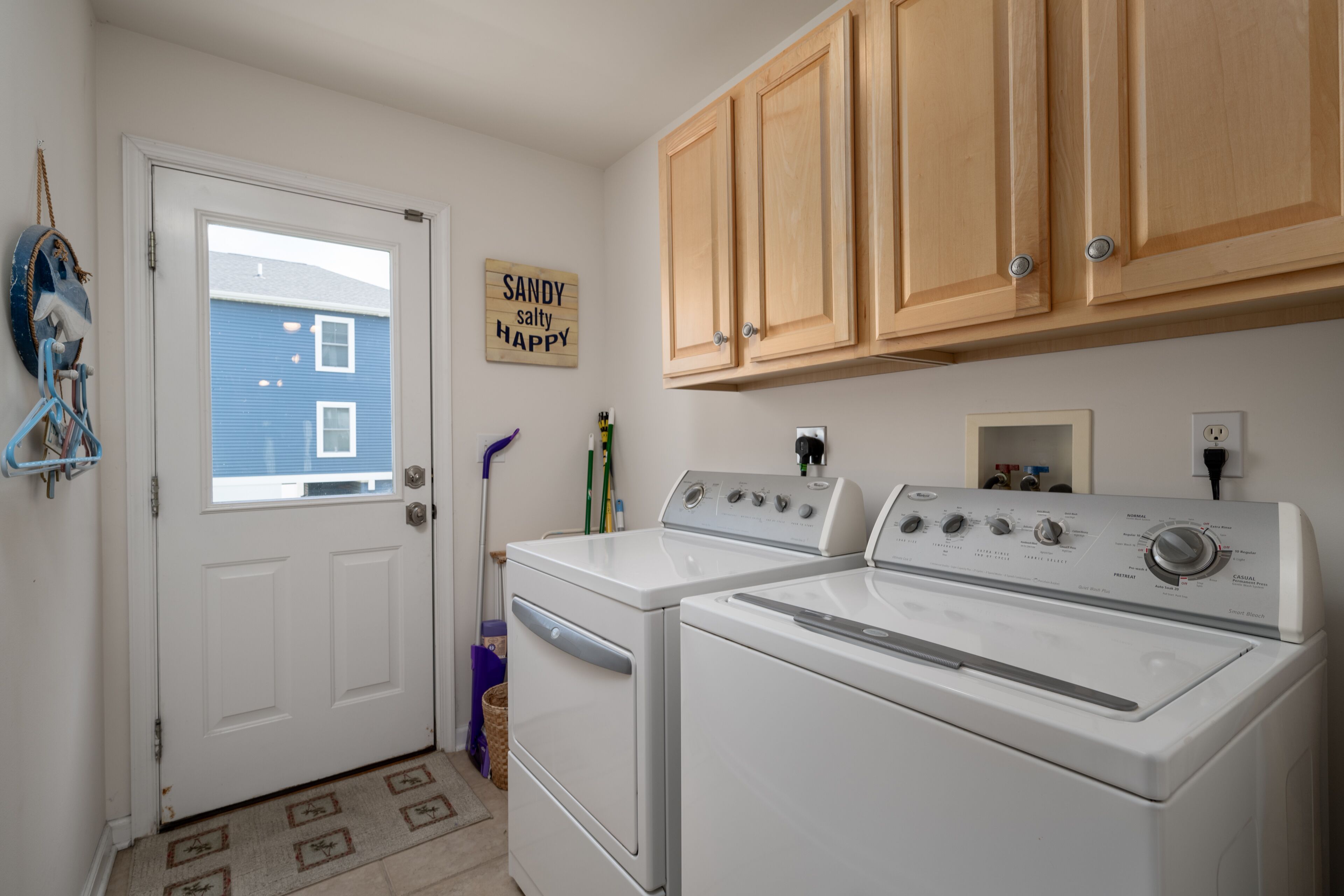 Une buanderie avec deux lave-linge et deux sèche-linge blancs, des armoires en bois, une porte donnant sur une maison bleue, et une pancarte sur laquelle on peut lire : « SANDY, salé, heureux ».