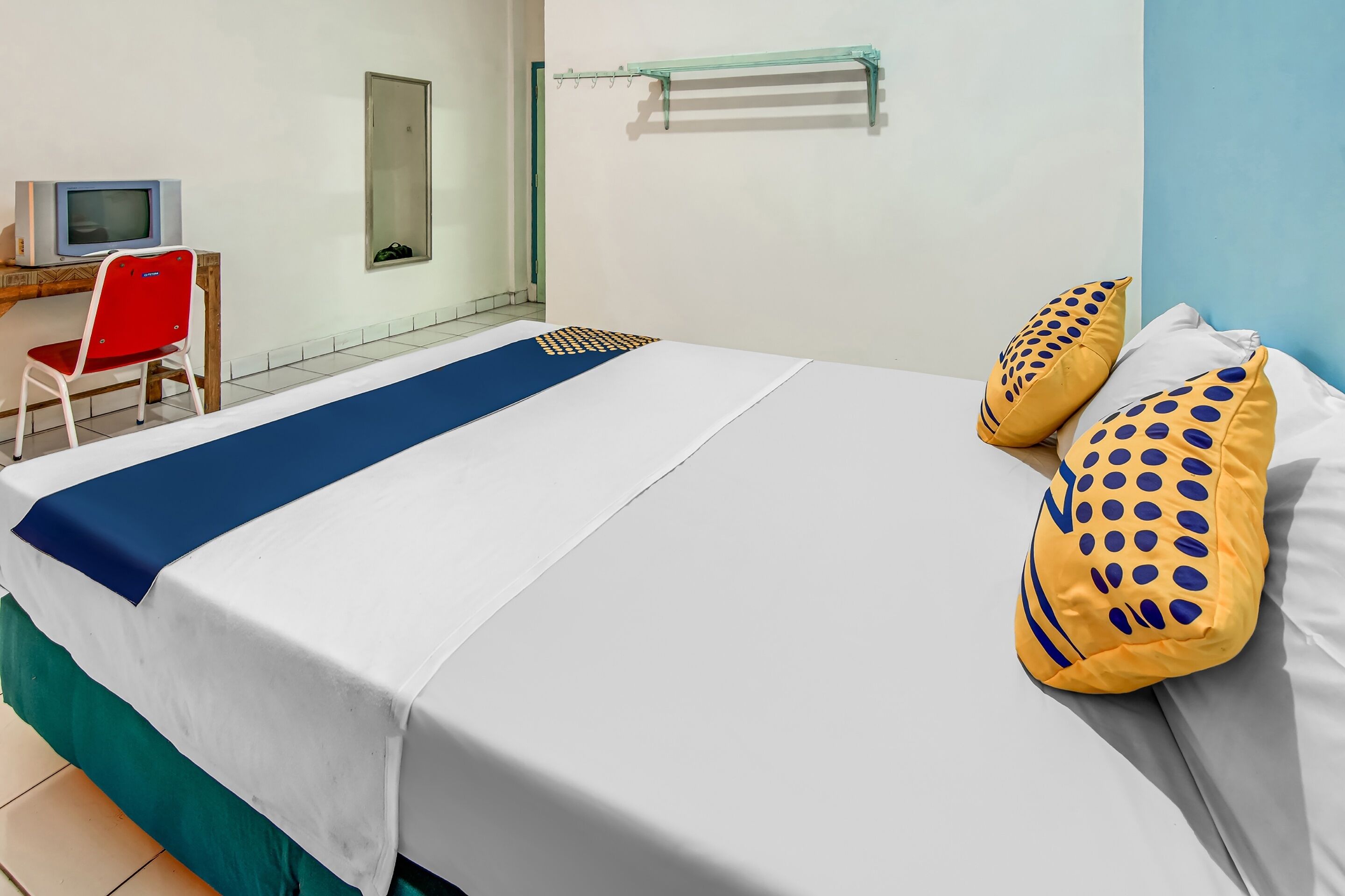 A neatly made bed with a blue and white bedspread, two yellow pillows with blue dots, a small desk with a television, and a red chair.