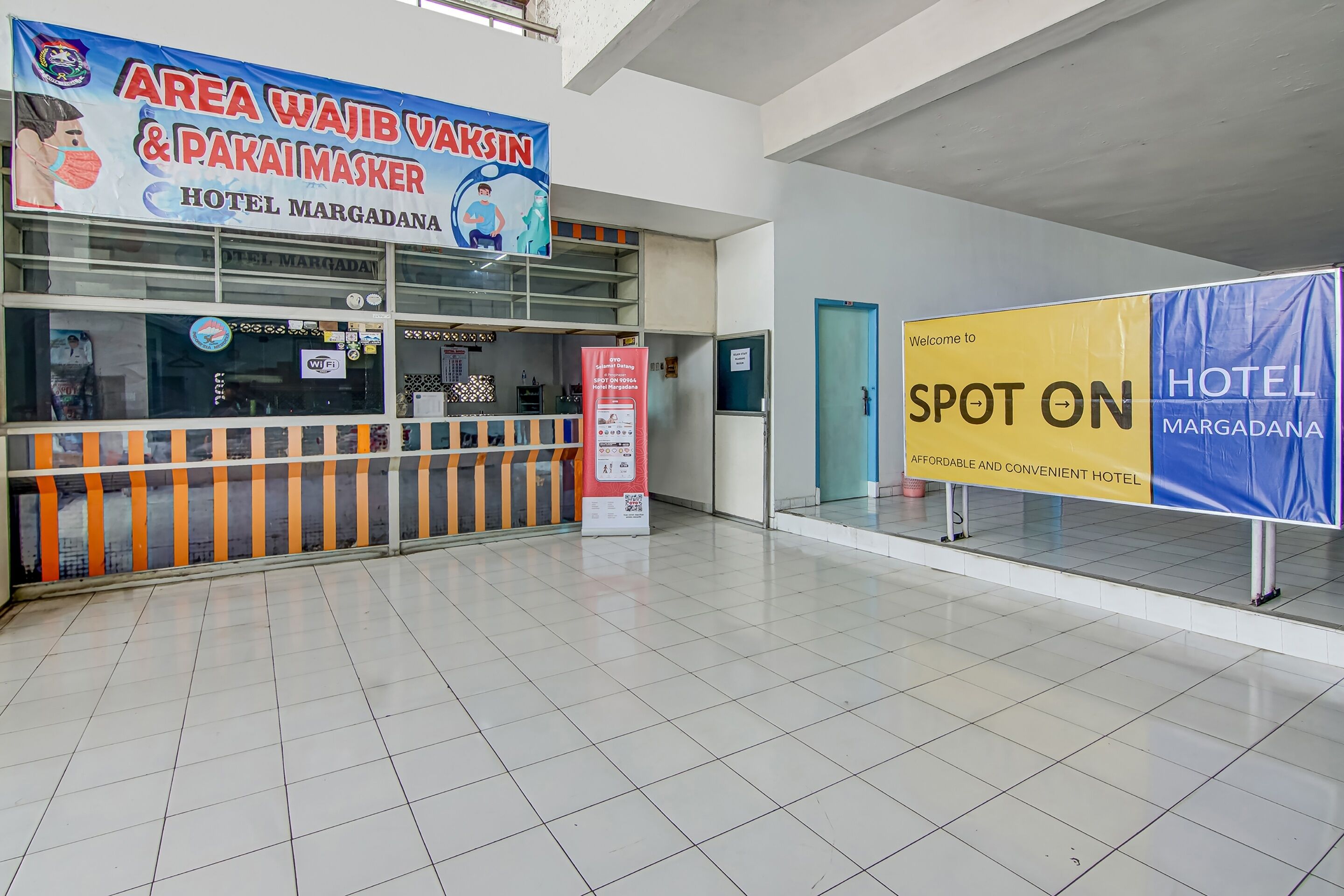 A hotel lobby with a reception desk, a sign for 'Area Wajib Vakkin & Pakai Masker', and a 'Spot On Hotel Margadana' sign.