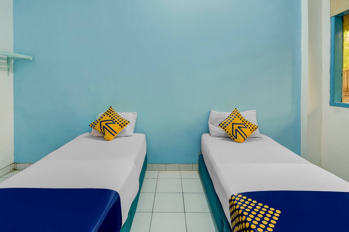 Une chambre avec deux lits simples, chacun doté d’un matelas blanc et d’une couvre-lit bleue. Chaque lit est agrémenté d’oreillers jaunes à motifs géométriques. Les murs sont peints en bleu clair, et une fenêtre à cadre bleu se trouve sur le côté droit.