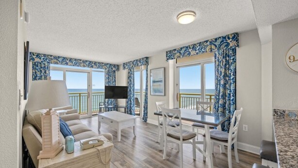 A spacious living room with a large window, a flat-screen TV, a dining table with chairs, and a sofa. - Bay Watch 1640-2BR Oceanfront - Corner Unit (North Myrtle Beach)