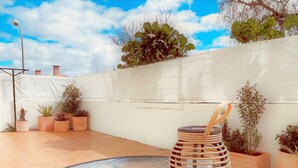 Une table dont le plateau est en verre, avec un panier tressé au centre, entourée de plantes en pot et d’un mur blanc percé d’une fenêtre.