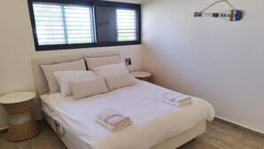 A neatly made bed with white linens and two folded towels, a bedside table with a lamp, a wicker basket, and a wall-mounted shelf with a decorative item. - YalaRent Cliff side villa - private pool (Eilat)