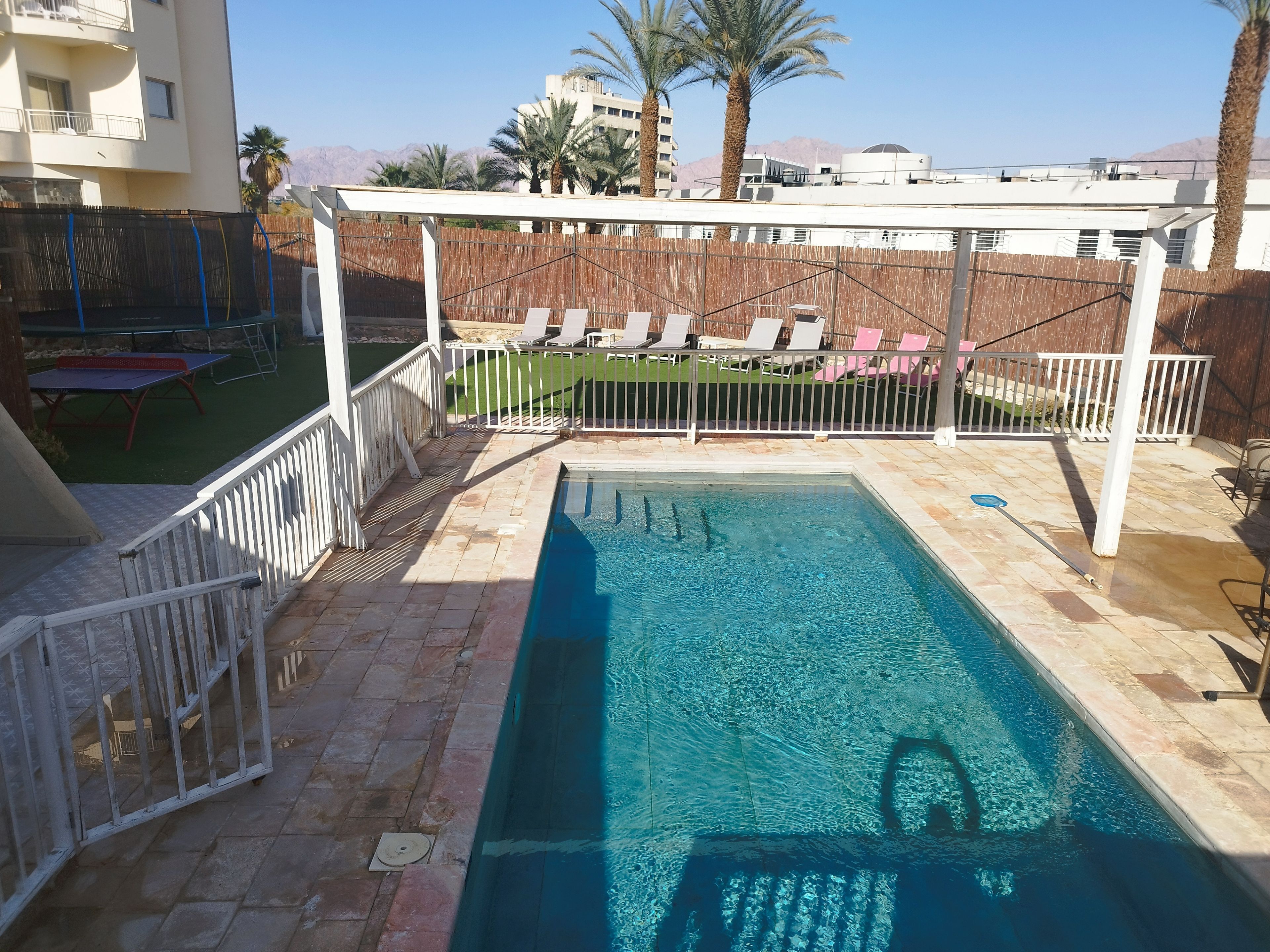 A swimming pool with a pergola and lounge chairs, surrounded by a fenced patio area with palm trees and a mountain view in the background.