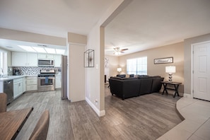 A modern kitchen with white cabinets, a microwave, and an oven. There is a dining area with a wooden table and chairs. The living room features a black sofa and a lamp.