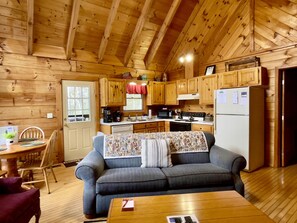 Interior acolhedor de uma cabana com teto de madeira, cozinha compacta, sofá e área de refeições.