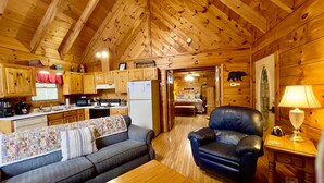 A cozy cabin interior with a wooden ceiling, a kitchenette, a living room with a sofa and armchair, and a bedroom visible through an open door.
