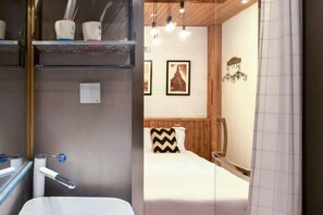 A modern bathroom with a white sink, a mirror, and a shower area. There are framed pictures on the wall and a bed with a chevron-patterned pillow in the background.