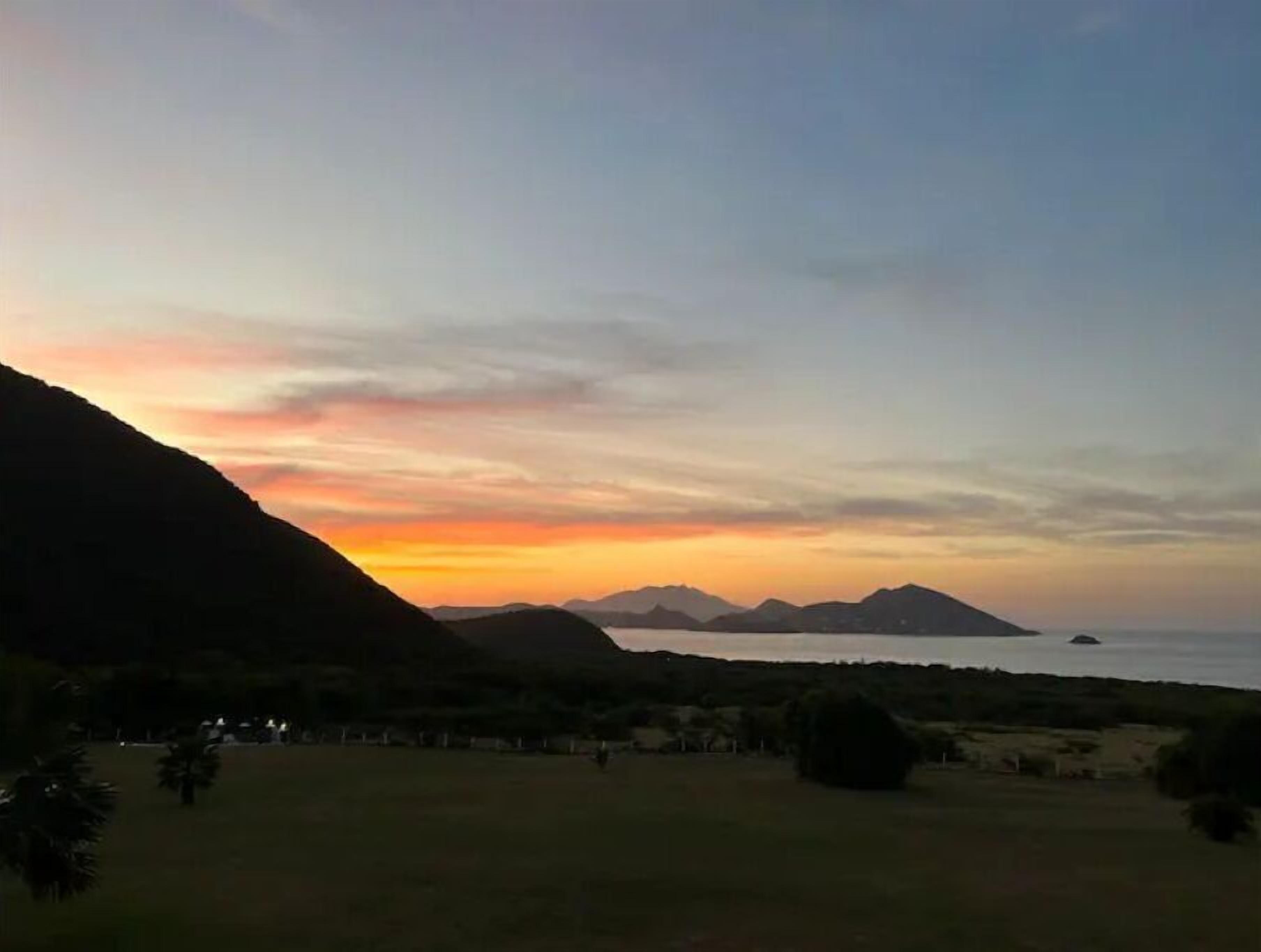 A coastal landscape with a sunset, mountains, and a grassy field.