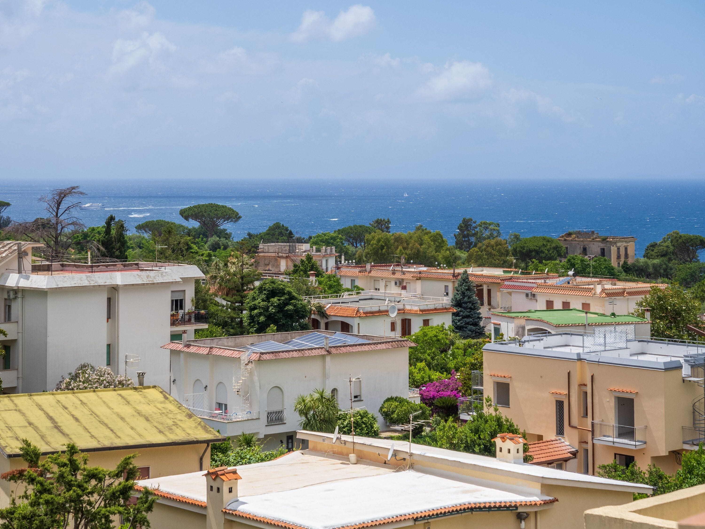 A coastal residential area with terracotta-tiled buildings, green spaces, and a view of the sea.