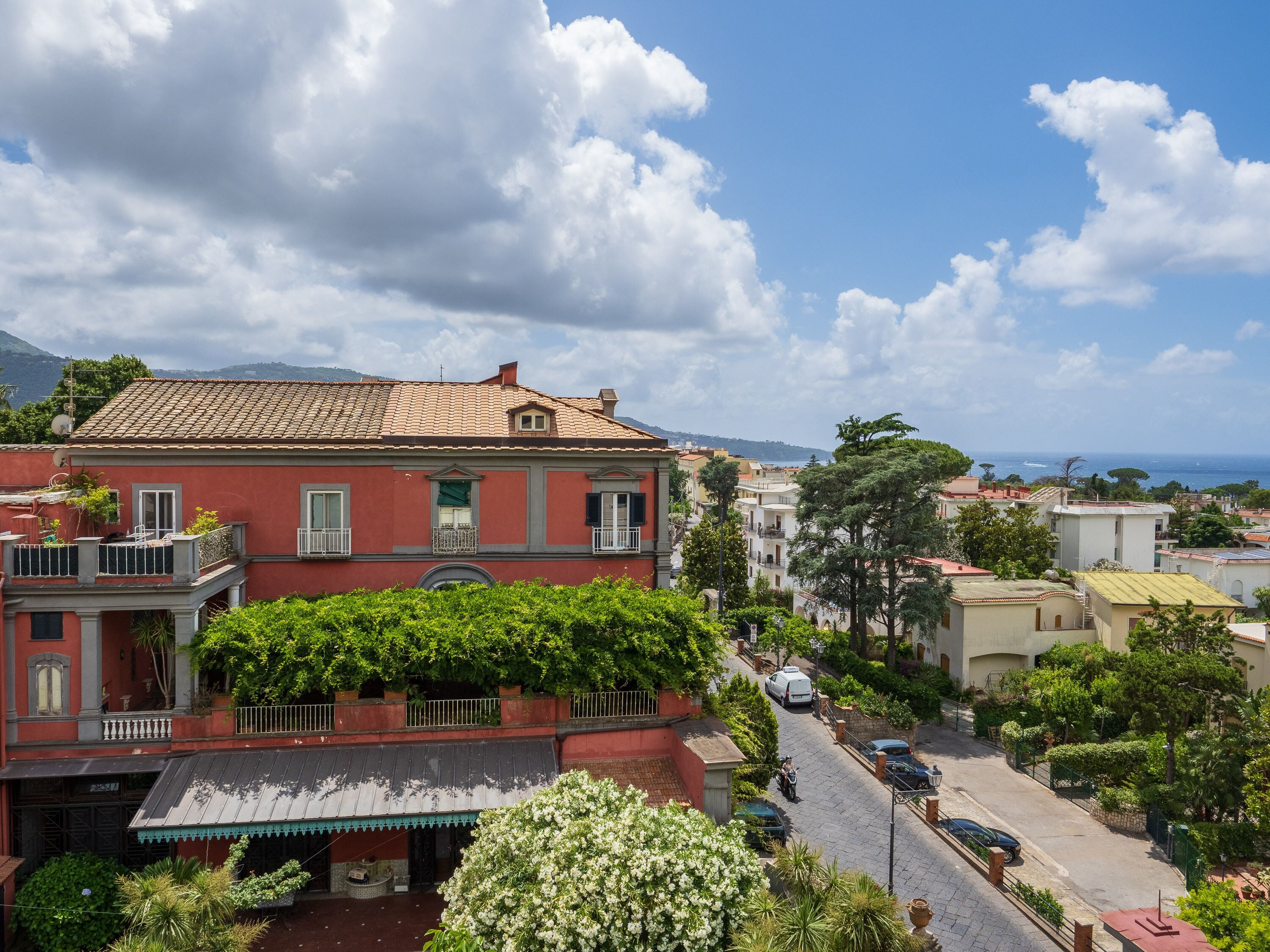 A red building with a tiled roof, a balcony with a railing, and a small garden area with white flowers.