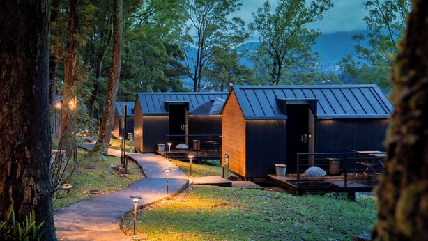 A row of cabins with metal roofs, surrounded by trees, and a paved path leading to them. - Bobocabin Coban Rondo Malang (Pujon)
