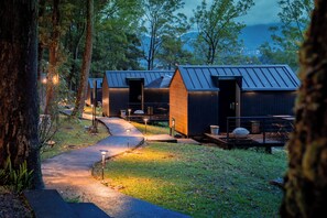 Une rangée de cabanes aux toits métalliques, entourées d’arbres, et un chemin pavé qui y mène.