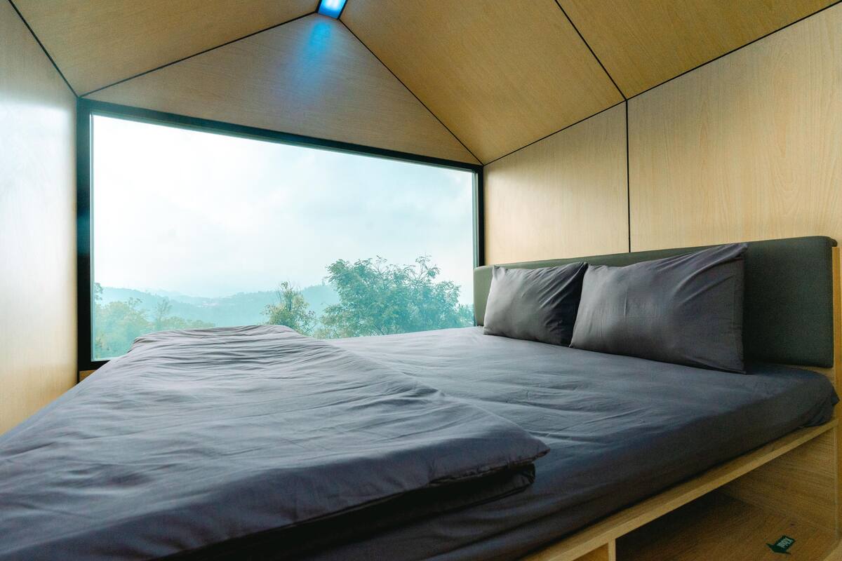 A modern bedroom with a large window, a bed with grey bedding, and wooden walls.