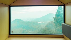 A room with a large window offering a view of a forested valley and distant hills. - Bobocabin Coban Rondo Malang (Pujon)