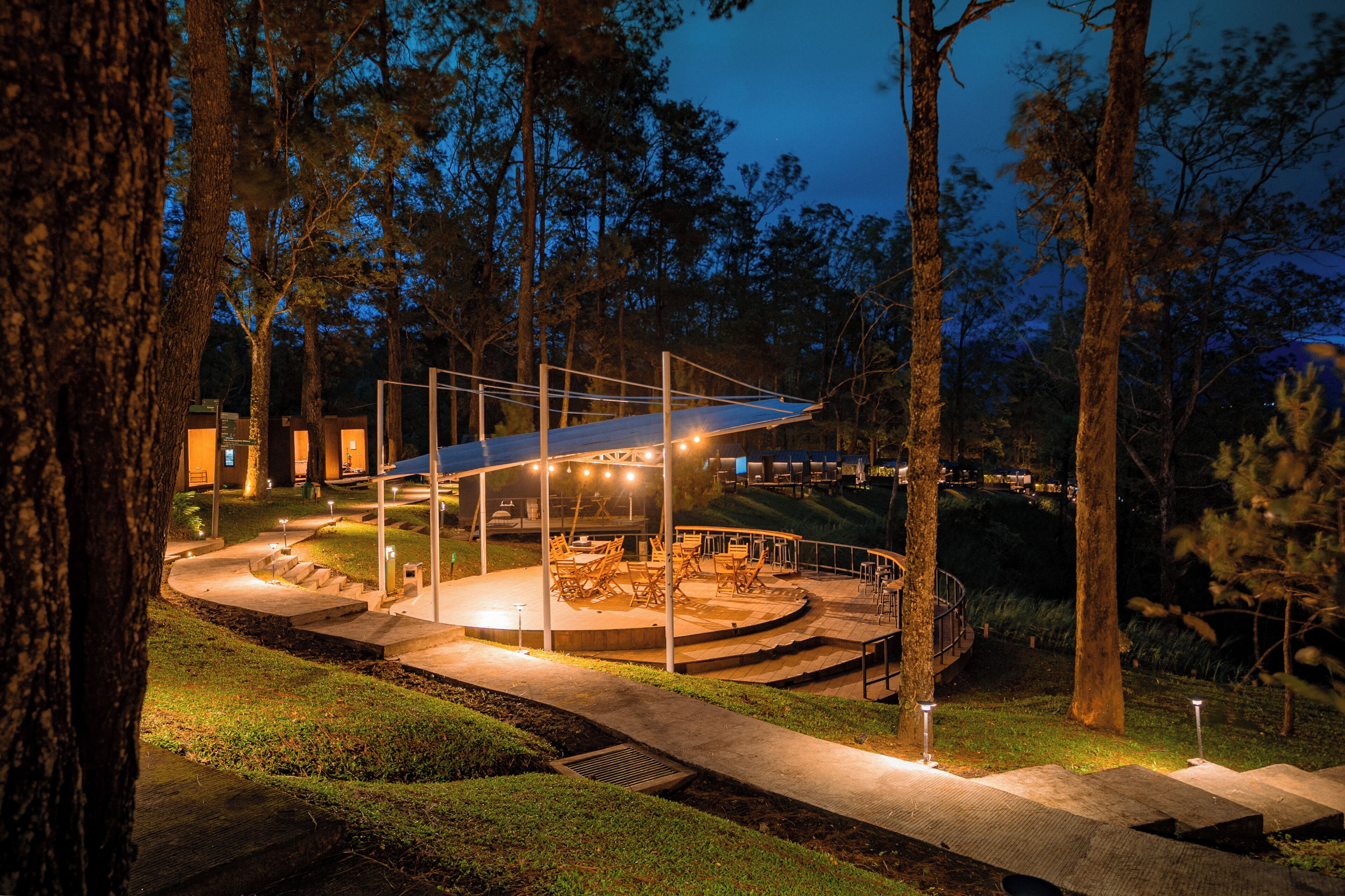 A covered outdoor seating area with tables and chairs, surrounded by trees and a well-lit pathway.