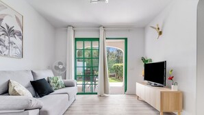 Una sala moderna con un sofá gris, un mueble de televisión de madera y un ventanal con contraventanas verdes.