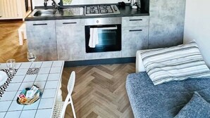 A compact kitchen with grey cabinets, a stove, and a sink. There's a dining table with a white chair and a grey sofa with striped cushions. - one bedroom apartment with parking, 5 minutes walk from piazzale Arnaldo (Brescia)