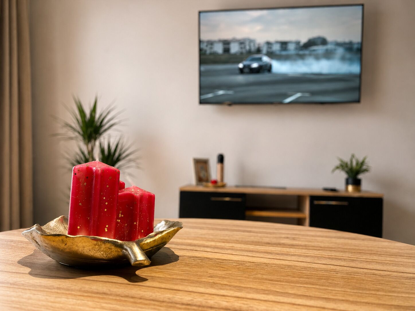 Ein Holztisch mit einer dekorativen Schale, die ein rotes Objekt enthält, ein an der Wand befestigter Fernseher und eine Pflanze im Hintergrund.