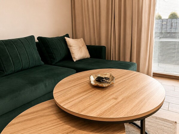 Un canapé vert avec un coussin beige, une table basse en bois surmontée d’un bol décoratif et une fenêtre avec des stores.