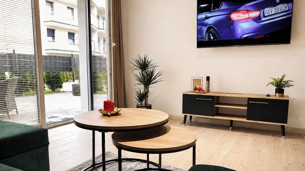 A modern living room with a round wooden table, a black TV on the wall, and a view of a building outside.