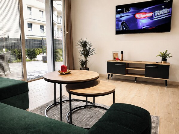 A modern living room with a round wooden table, a black TV on the wall, and a view of a building outside.